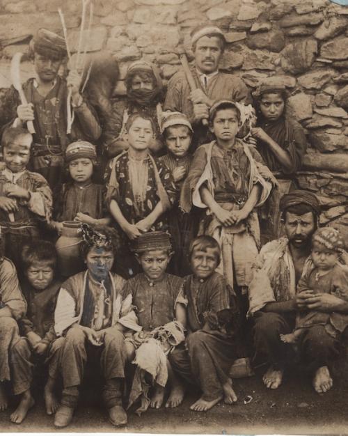 Historic photo of about 20 people, many of them children arranged in rows against a stone wall of a building